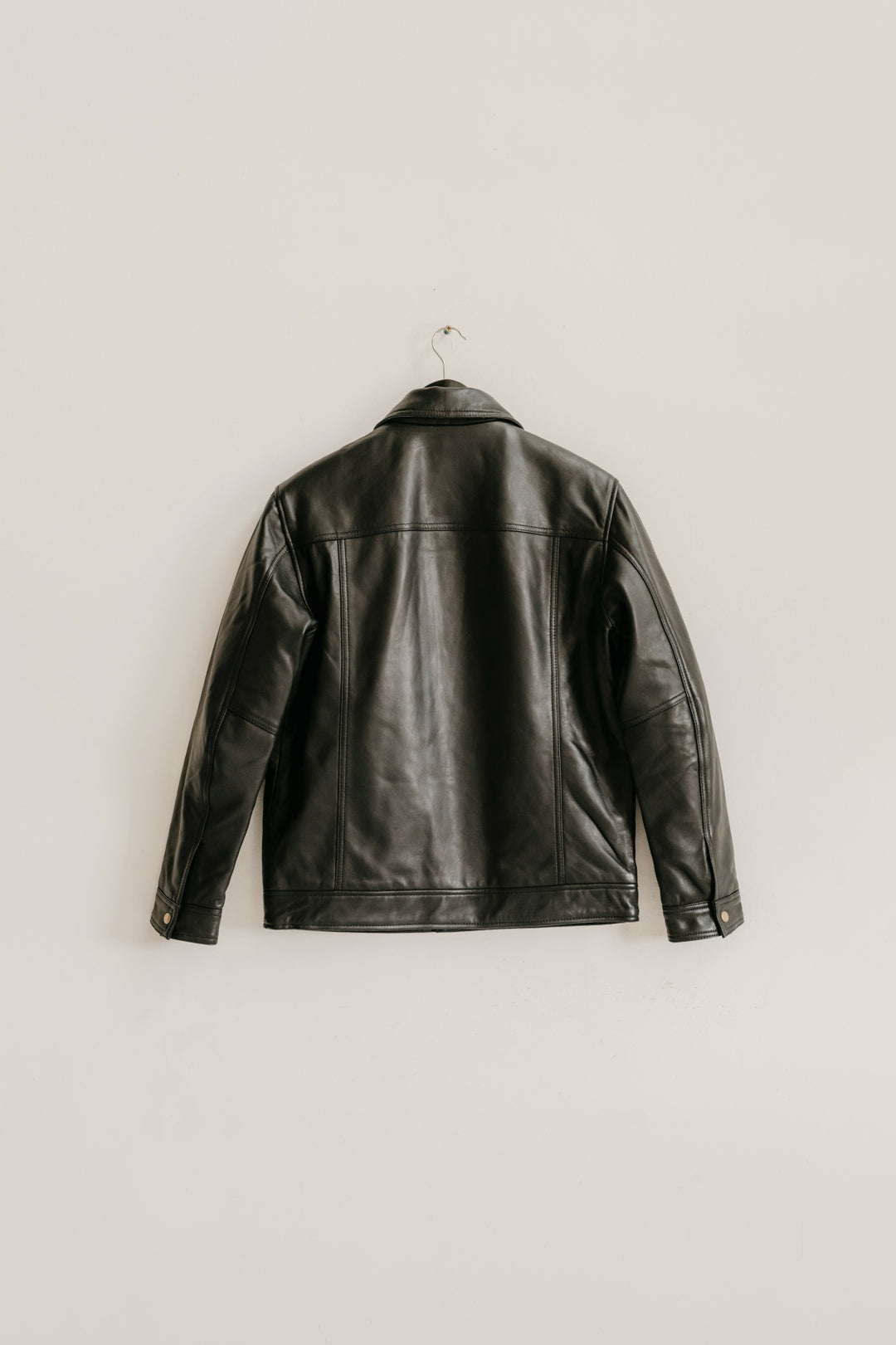 Back view of the Hallwood Black Leather Jacket by Bolt + Buckle hanging on a white wall.