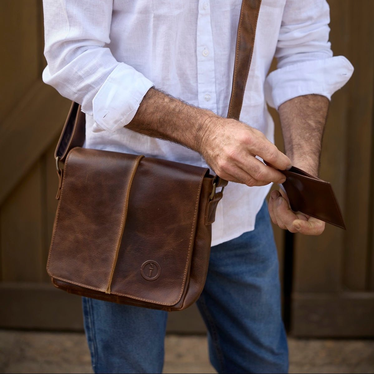 Man holding a brown leather bag and wallet against a wooden background