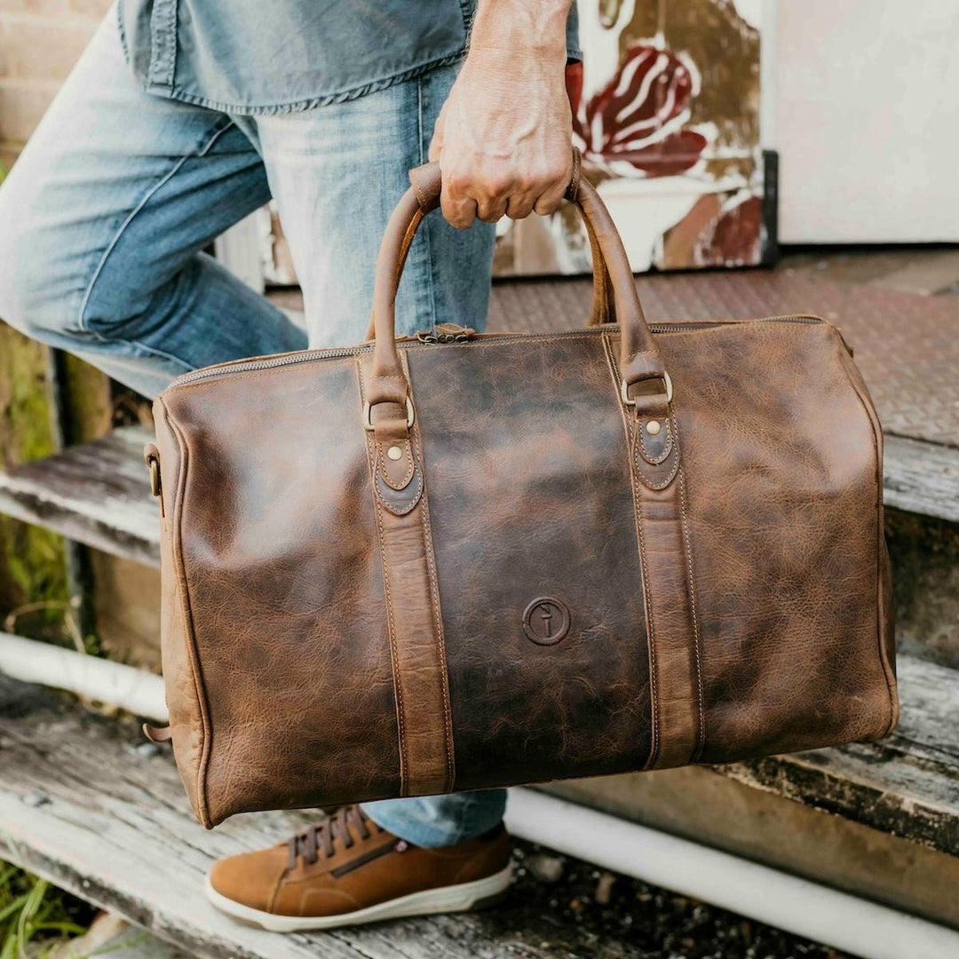 Person holding a brown leather duffel bag on wooden steps