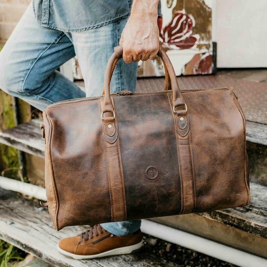 Person holding a brown leather duffel bag on wooden steps