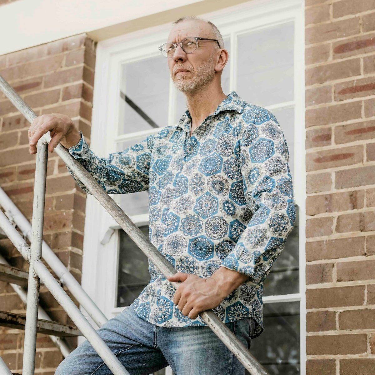 Man in a patterned shirt and jeans standing on a staircase against a brick wall.