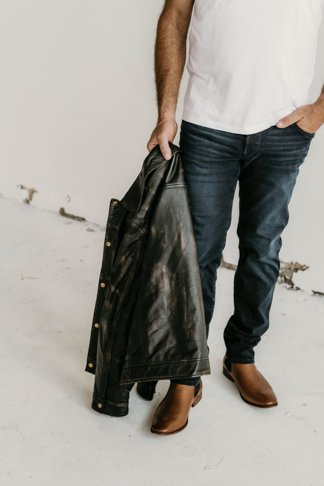 Close-up of man holding the Hailwood Distressed Brown Leather Jacket by Indepal Leather from Bolt + Buckle, styled with dark jeans and brown boots.