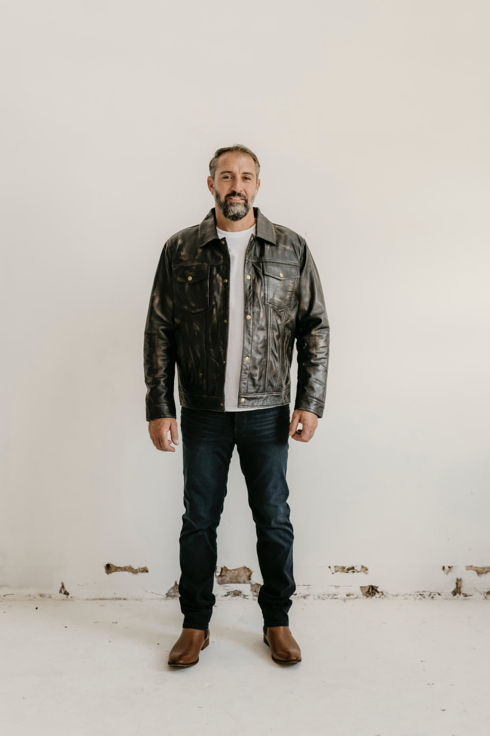 Man wearing the Hailwood Distressed Brown Leather Jacket by Bolt + Buckle, styled with dark jeans and brown boots, standing against a white wall.