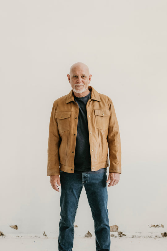 Man wearing the Hailwood Raw Beige Leather Jacket by Indepal Leather from Bolt + Buckle, styled with a black t-shirt and dark jeans, standing against a white wall.