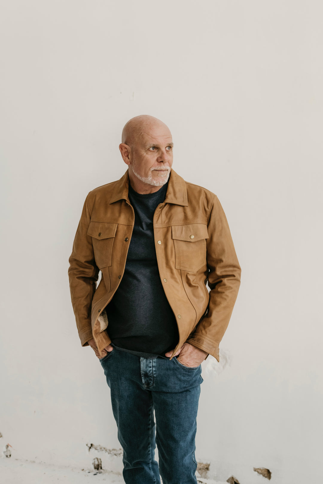 Man wearing the Hailwood Raw Beige Leather Jacket by Indepal Leather from Bolt + Buckle, styled with a dark t-shirt and jeans, standing against a white wall with hands in pockets.