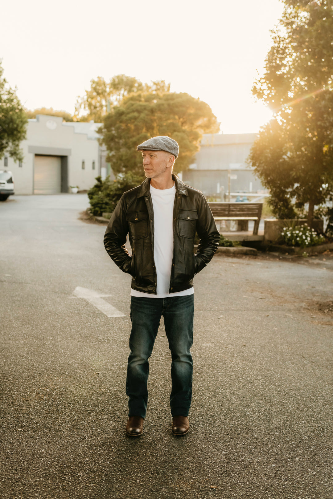 Man wearing the Hallwood Black Leather Jacket by Bolt + Buckle, styled with jeans and a flat cap in a sunlit street setting.