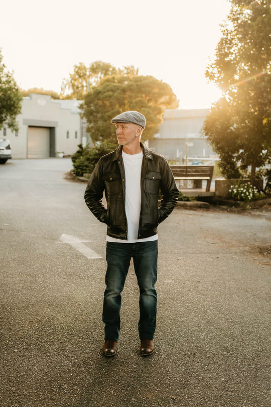Man wearing the Hallwood Black Leather Jacket by Bolt + Buckle, styled with jeans and a flat cap in a sunlit street setting.