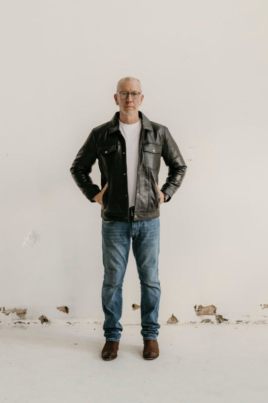 Man wearing the Hallwood Black Leather Jacket by Bolt + Buckle, styled with jeans and boots, standing against a white wall.