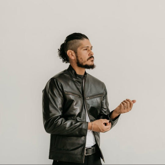 Man wearing the Ollie Black Leather Jacket by Indepal Leather from Bolt + Buckle, adjusting his sleeve while standing against a white wall.