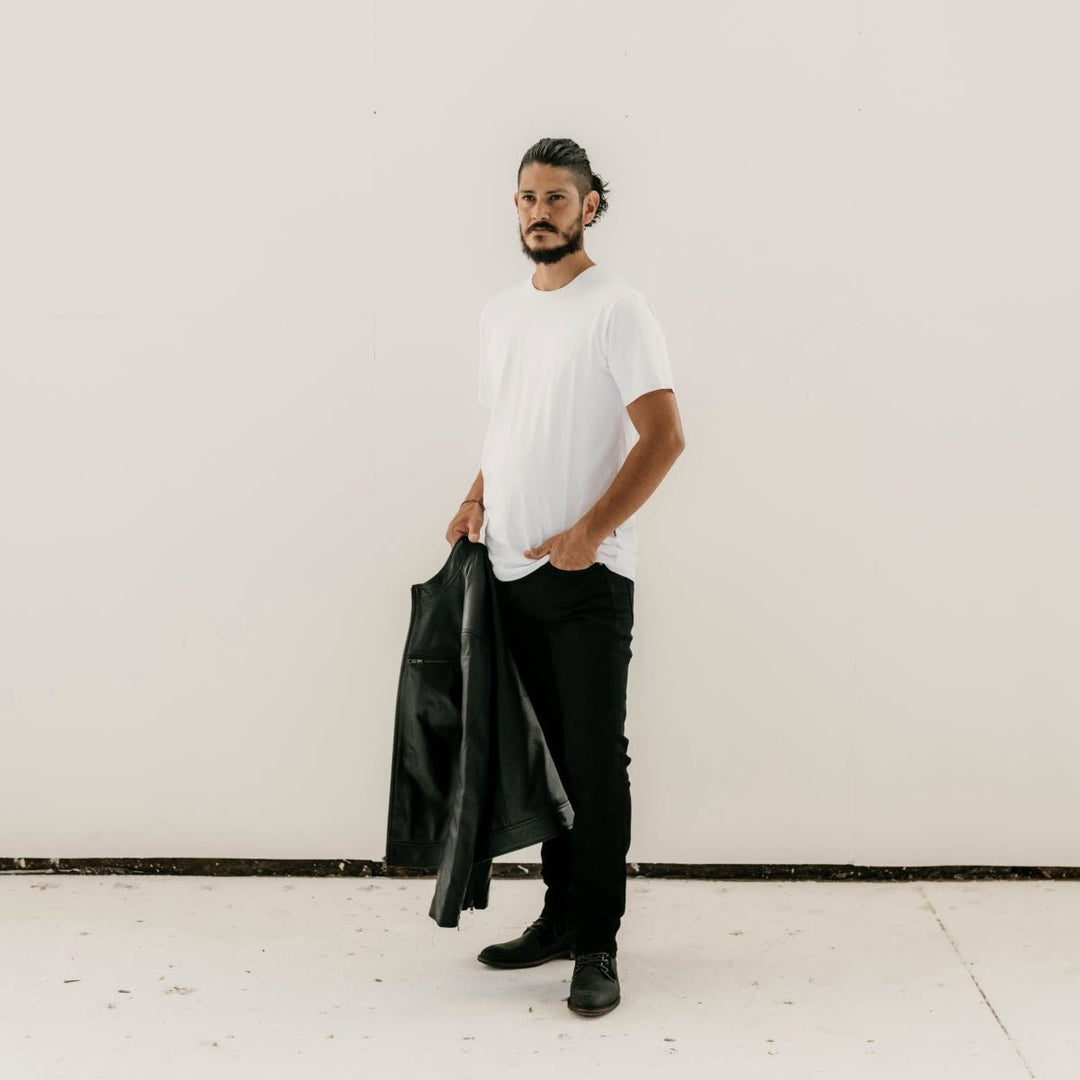 Man holding the Ollie Black Leather Jacket by Indepal Leather from Bolt + Buckle, styled with a white t-shirt, black pants, and boots, standing against a white wall.