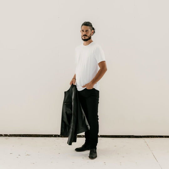 Man holding the Ollie Black Leather Jacket by Indepal Leather from Bolt + Buckle, styled with a white t-shirt, black pants, and boots, standing against a white wall.