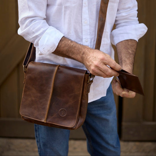 Man holding a brown leather bag and wallet against a wooden background